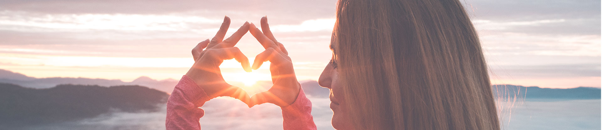 person holding hands in heart shape with sunset, clouds and hills in the background