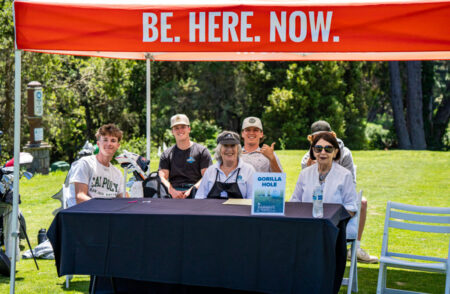 group of people smiling and sitting under a banner that reads be here now