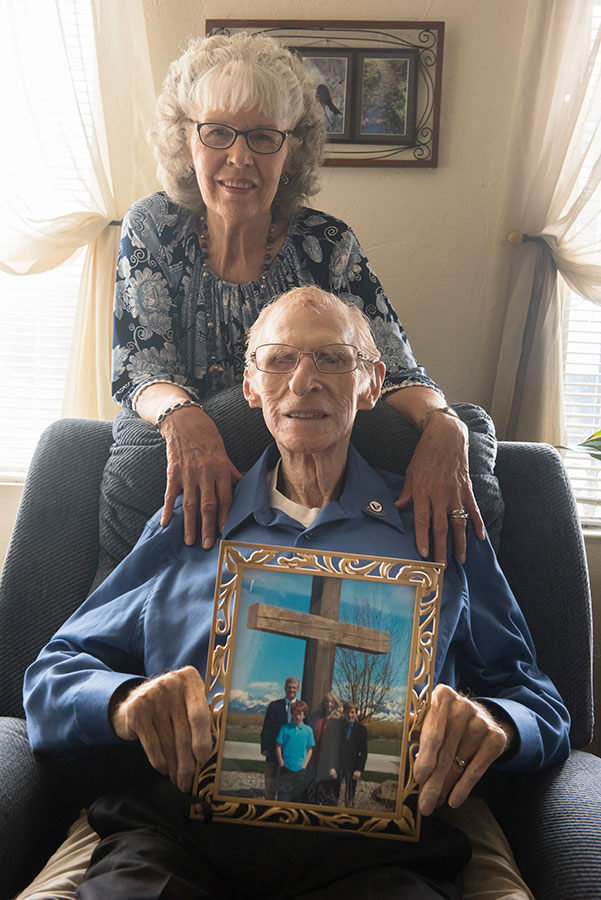 elderly couple with the husband holding a framed picture of a family in front of a big cross